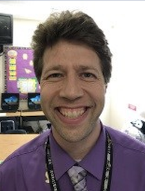 An online student and civics teacher in a classroom with a purple shirt.
